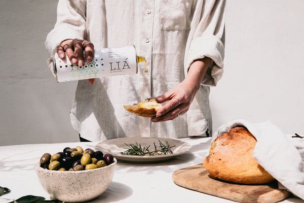 gedekte tafel met daarop een schaal olijven, een bord met een kruid en daarboven houdt een vrouw een brood waarop ze de olijfolie schenkt